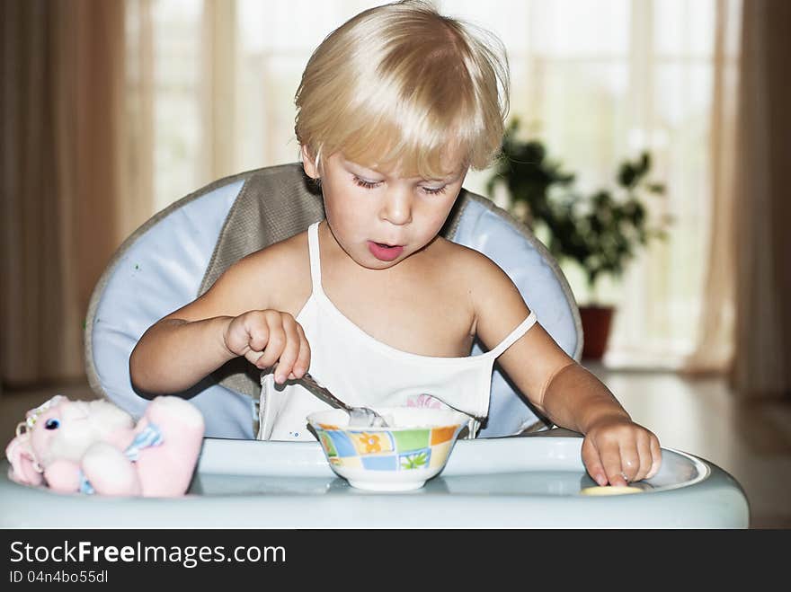 The nice girl (2 years), the blonde, sits at a little table and eats porridge. The nice girl (2 years), the blonde, sits at a little table and eats porridge