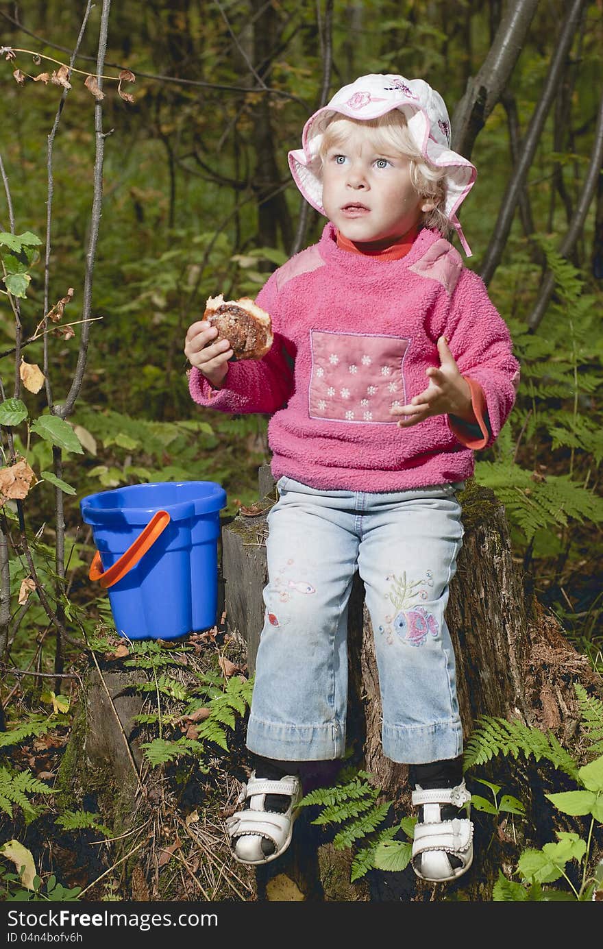 The nice girl (2 age), the blonde, sits on hemp in the wood and eats a roll. The nice girl (2 age), the blonde, sits on hemp in the wood and eats a roll