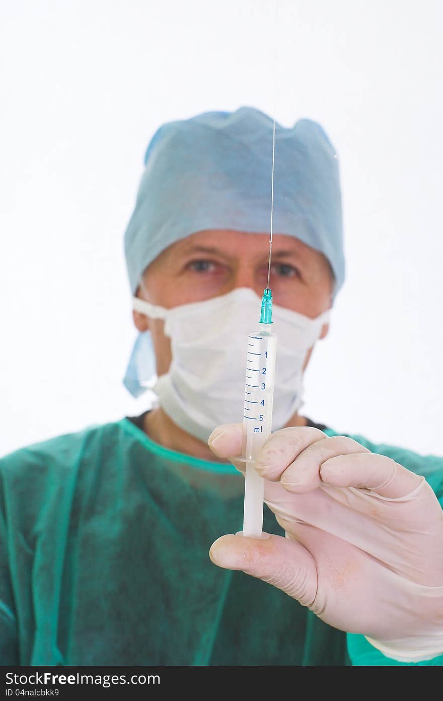 Portrait of a surgeon preparings an injection. He keeps the full syringe the needle up, removing a bubble of air. He wears rubber gloves, a mask, a cap and a green surgical coat. Portrait of a surgeon preparings an injection. He keeps the full syringe the needle up, removing a bubble of air. He wears rubber gloves, a mask, a cap and a green surgical coat.