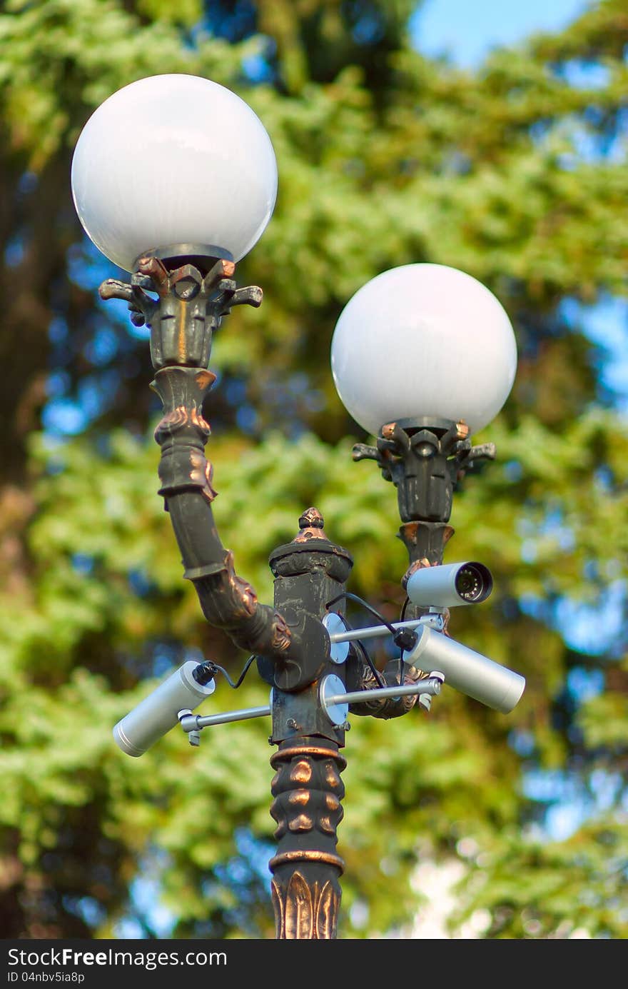 Security surveillance camera on a column with a lamp, on green tree background, selective focus