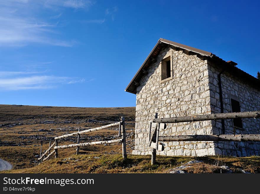 Old cabin in the mountain with stone walls, wooden fence in the green meadow and blue sky with clouds