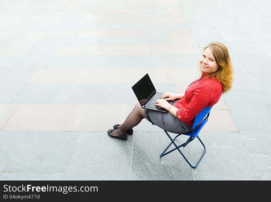 Image of the businesswoman sitting with the laptop and looking at the camera in the empty street. Focus is made on top of the background marbled tile in the street. Image of the businesswoman sitting with the laptop and looking at the camera in the empty street. Focus is made on top of the background marbled tile in the street.