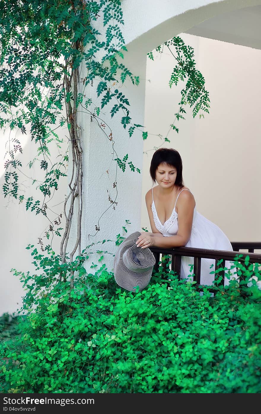 Girl in a white dress with black hat standing on the balcony and looking at a green tree. Girl in a white dress with black hat standing on the balcony and looking at a green tree