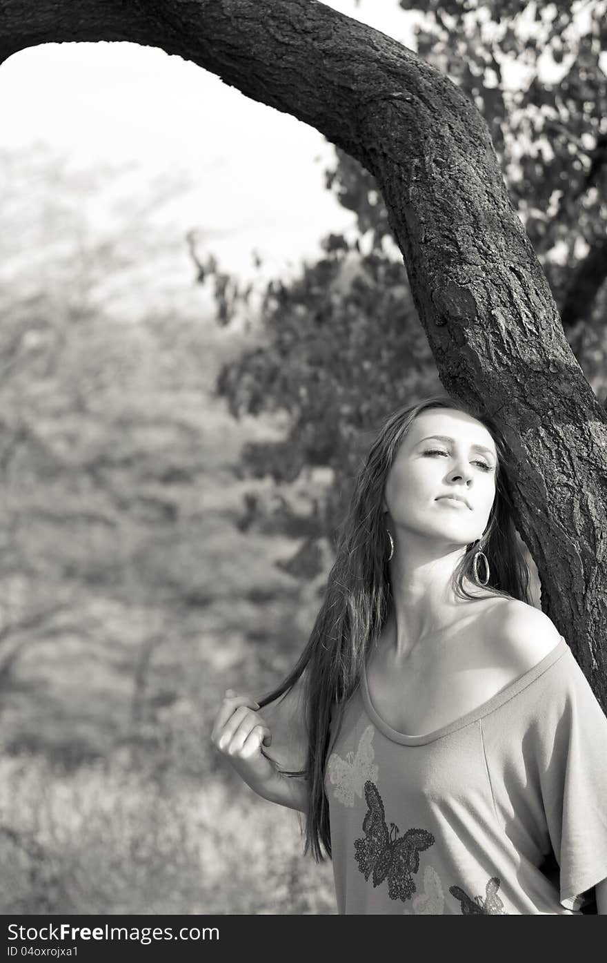 Black and white photograph of a pretty young brunette woman with long hair standing near a tree and enjoying the sun on a bright day. Black and white photograph of a pretty young brunette woman with long hair standing near a tree and enjoying the sun on a bright day