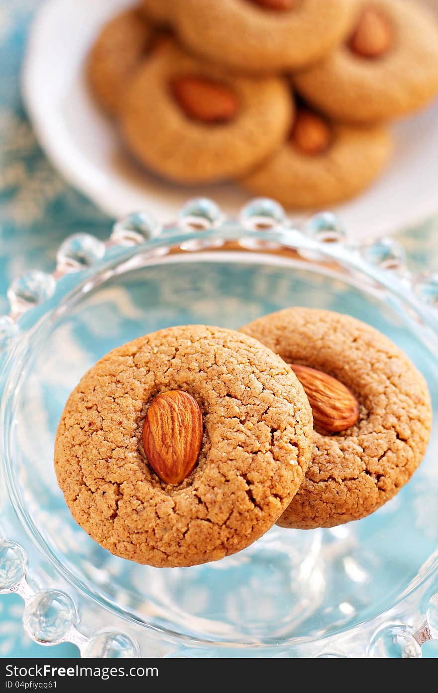 Two almond whole wheat cookies on a small glass cake stand with more cookies in the background