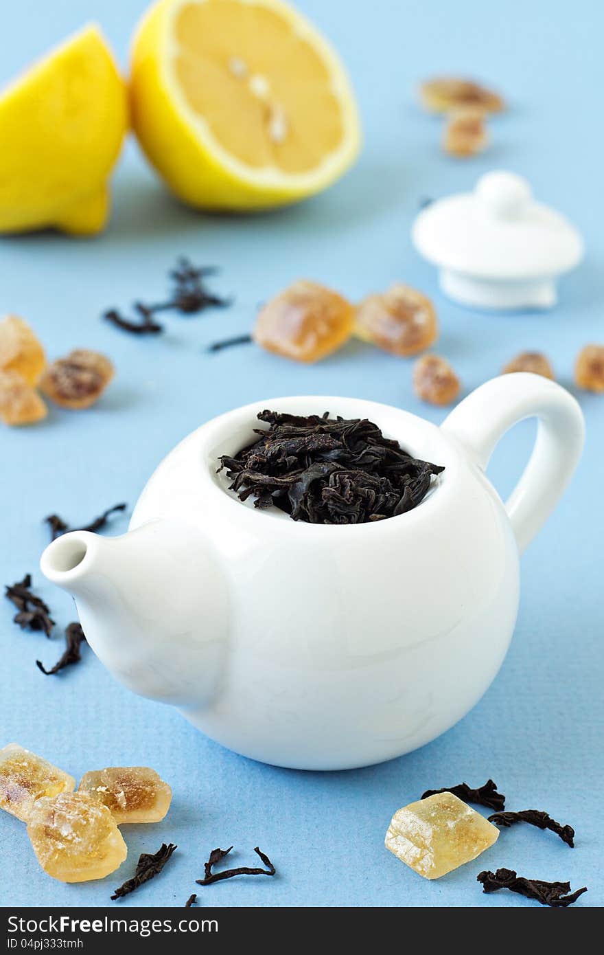 Small tea pot with leaves of black tea, brown crystal sugar and lemons on light blue background