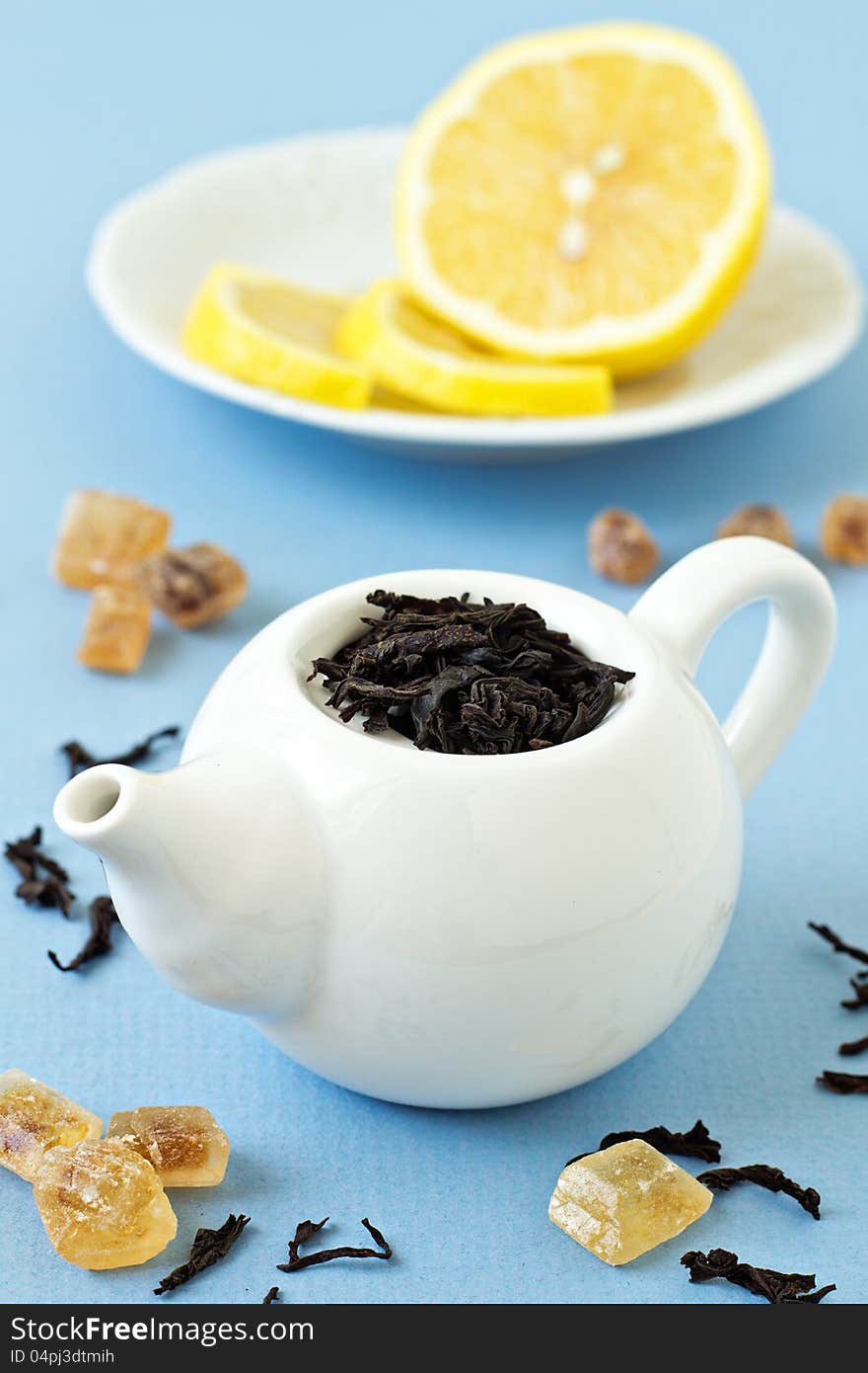 Small tea pot with leaves of black tea, brown crystal sugar and lemons on light blue background