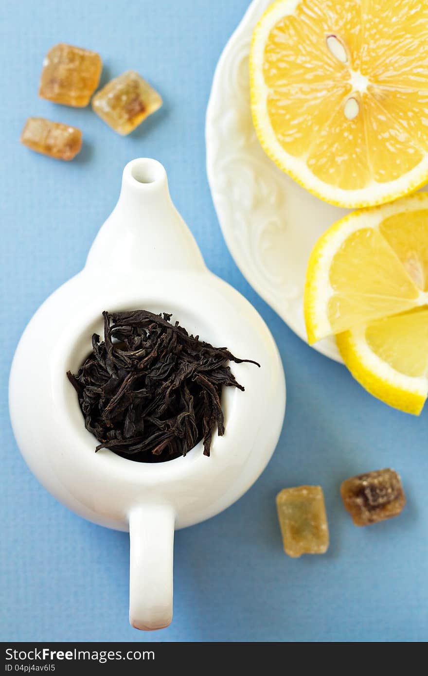 Small tea pot with leaves of black tea, brown crystal sugar and lemons on light blue background