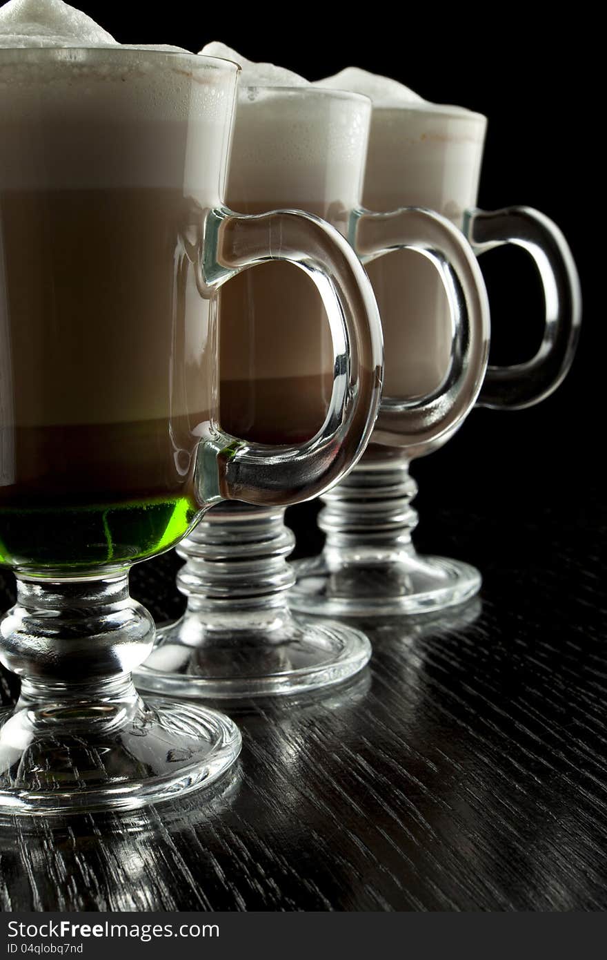 A group of three glasses of hot layered alcoholic cocktails, decorated with milk foam on black bar counter