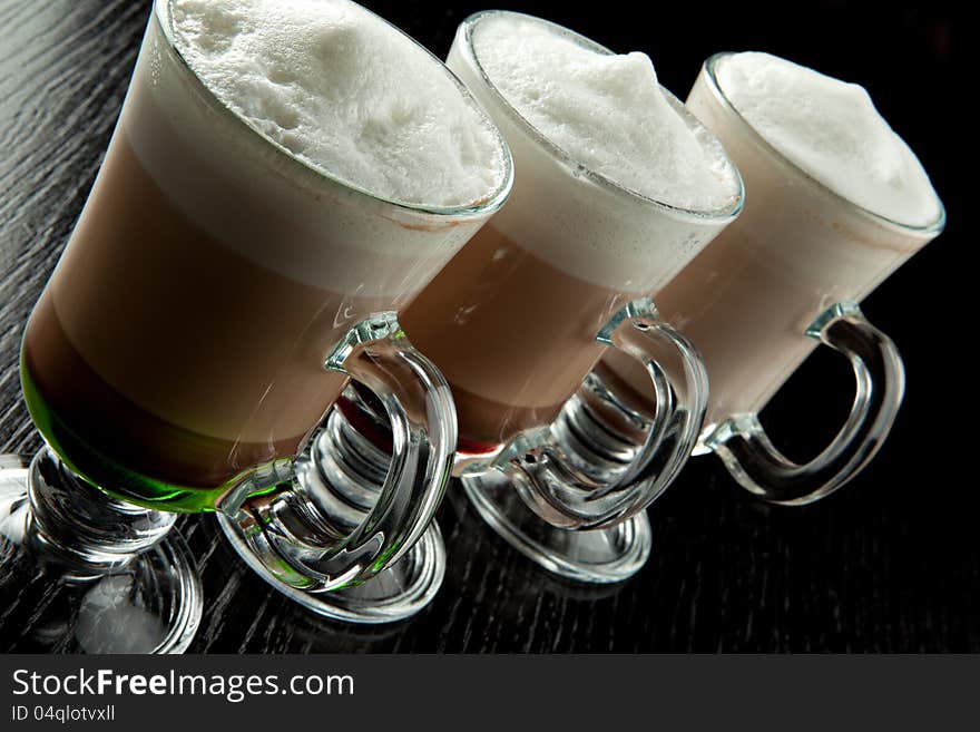 A group of three glasses of hot layered alcoholic cocktails, decorated with milk foam on black bar counter