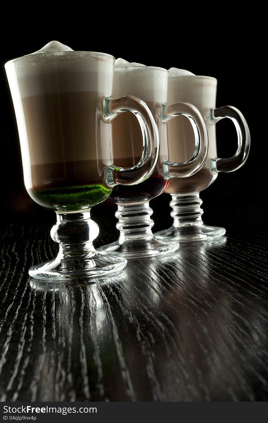 A group of three glasses of hot layered alcoholic cocktails, decorated with milk foam on black bar counter