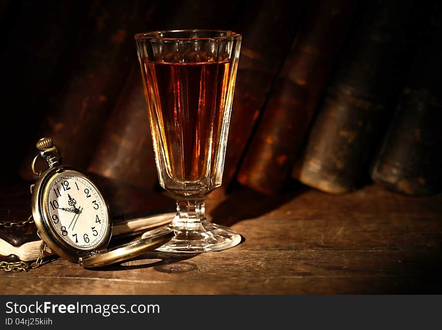 Vintage small wineglass with alcohol near open pocket watch on background with old books. Vintage small wineglass with alcohol near open pocket watch on background with old books