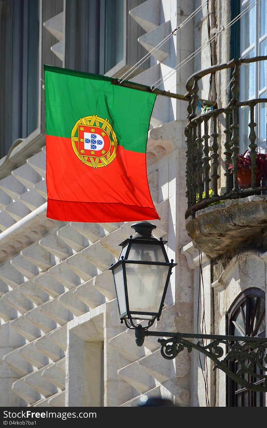 The flag of Portugal hangs from a house facade together with a street lamp and next to a balcony. The flag of Portugal hangs from a house facade together with a street lamp and next to a balcony.