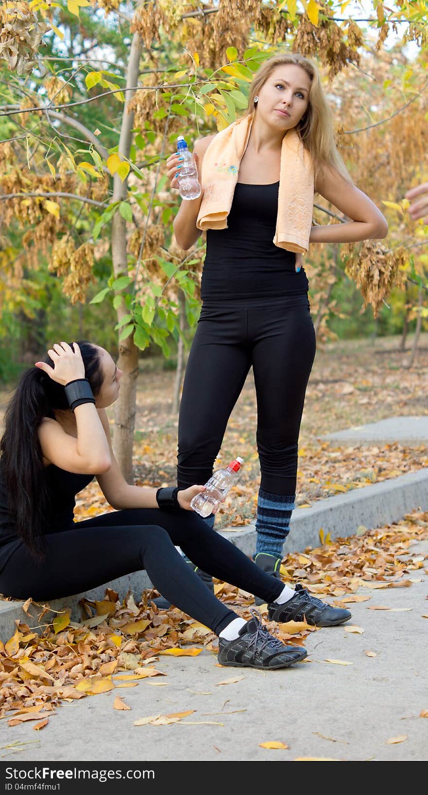 Two female athletes resting and drinking water sitting on the side of a rural road in their sportswear rehydrating after a heavy training session