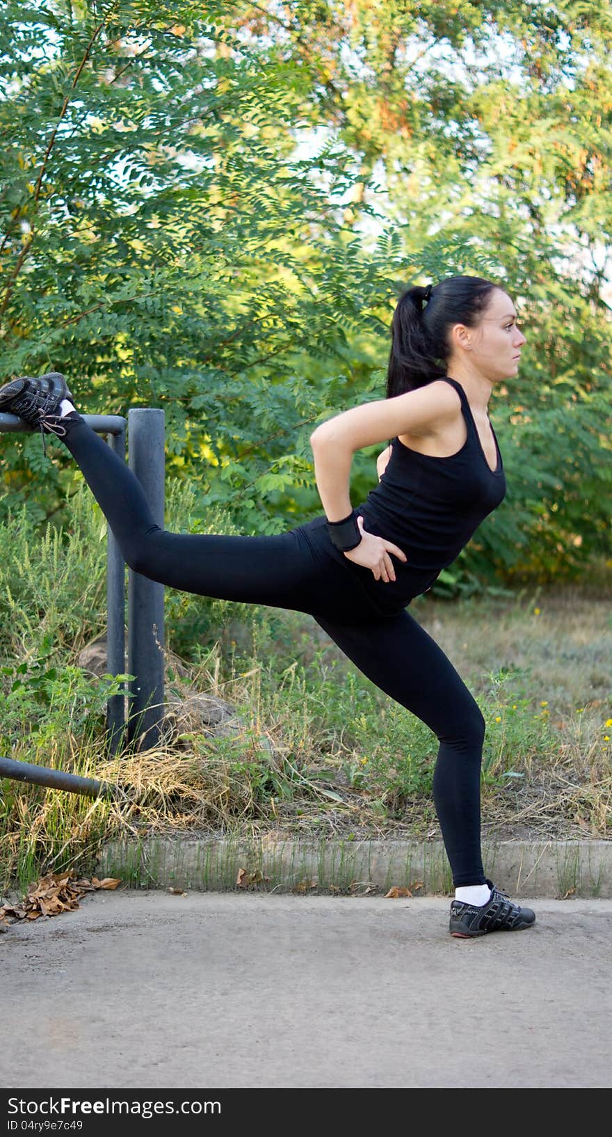 Fit healthy woman in sportswear doing exercises in the park using a metal railing to raised her leg for balance and control. Fit healthy woman in sportswear doing exercises in the park using a metal railing to raised her leg for balance and control