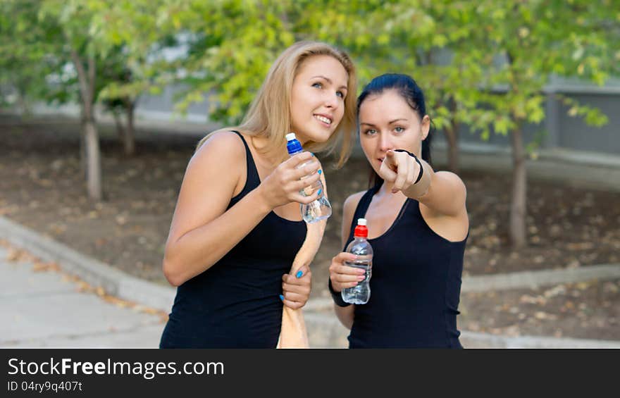 Woman athlete pointing out something to her team mate as they stand together chatting and drinking a refreshing bottle of water