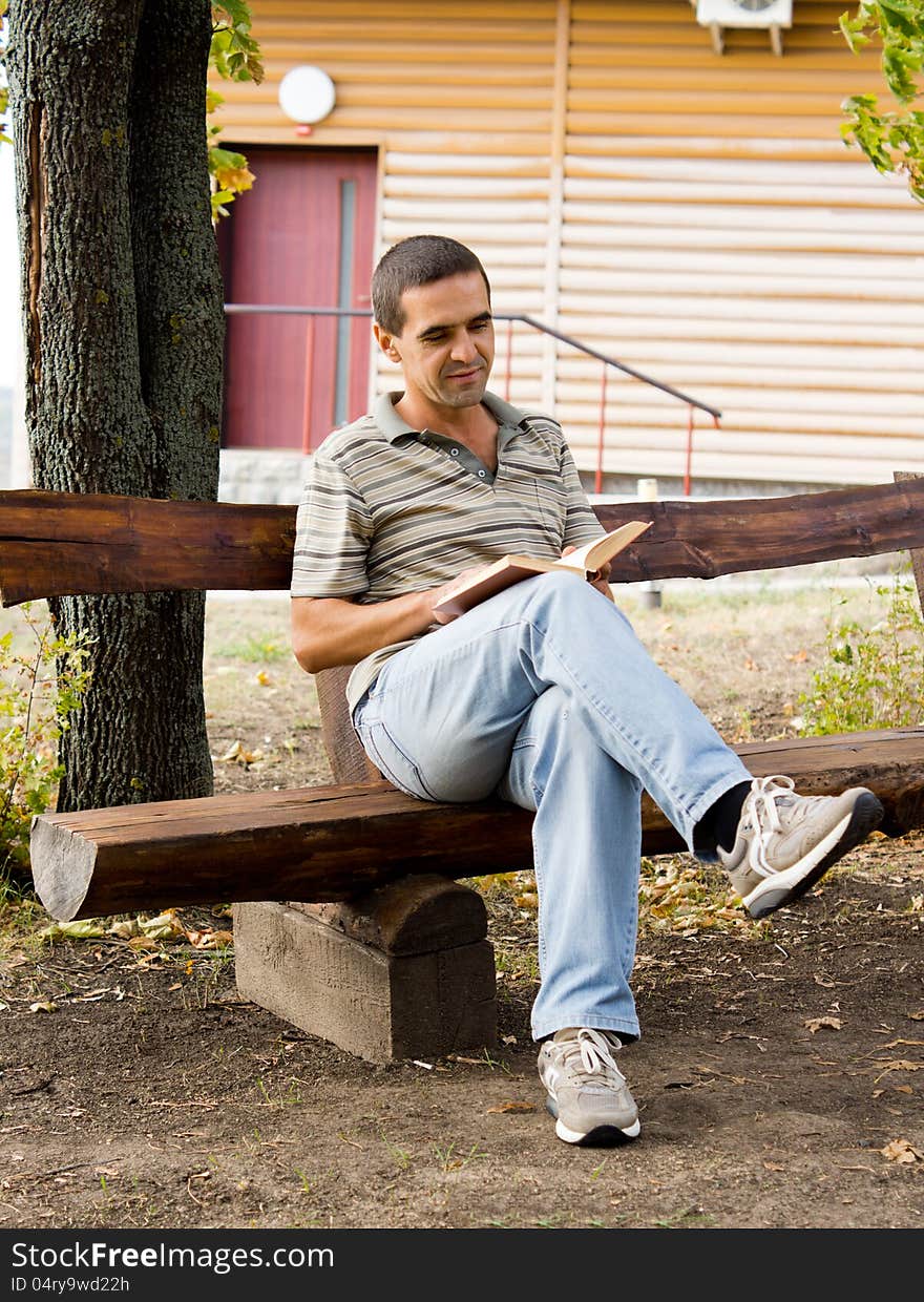 Man sitting relaxing reading a book on a rustic wooden bench in front of a timber log cabin. Man sitting relaxing reading a book on a rustic wooden bench in front of a timber log cabin