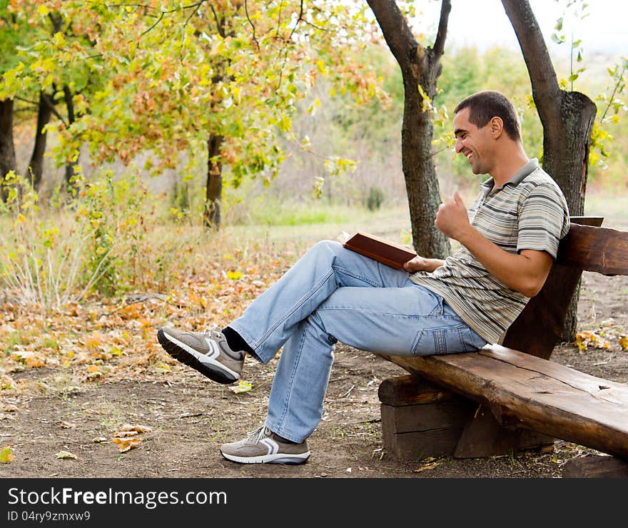 Middle-aged man sitting on a rustic wooden bench in the countryside laughing and reading as he thoroughly enjoys his book. Middle-aged man sitting on a rustic wooden bench in the countryside laughing and reading as he thoroughly enjoys his book.