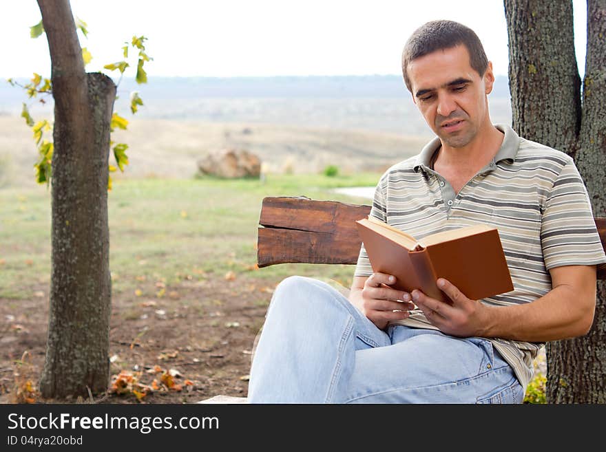 Middle-aged man sitting relaxing on a wooden bench enjoying a book in the garden with a view of open countryside and copyspace. Middle-aged man sitting relaxing on a wooden bench enjoying a book in the garden with a view of open countryside and copyspace