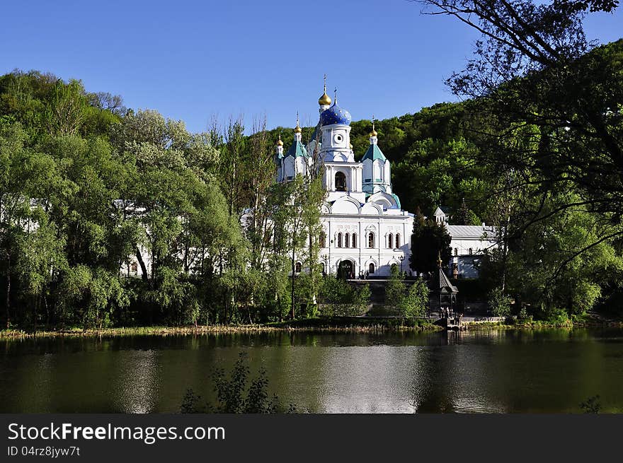 A temple at the river is surrounded by a green grove under blue sky