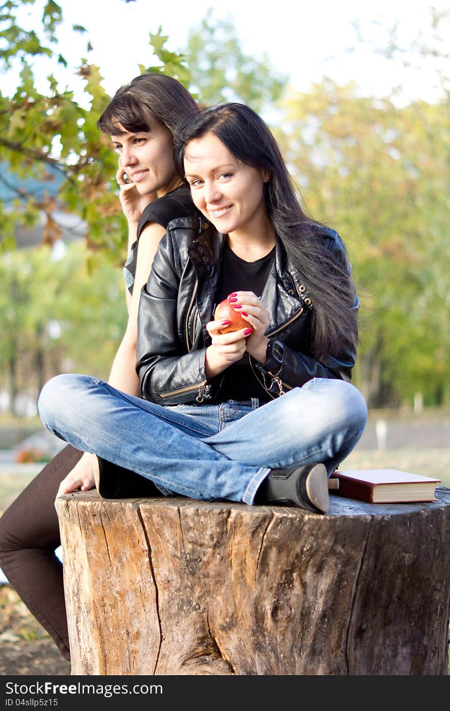 Young women seated on a sawn off tree trunk outdoors eating an apple while her friend chats on her mobile phone. Young women seated on a sawn off tree trunk outdoors eating an apple while her friend chats on her mobile phone