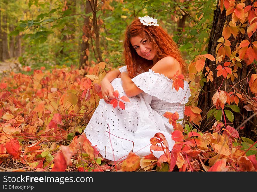 Beautiful red-haired young woman, wearing in white dress or the bride and decoration in form of flower on her head sitting among red leaves in autumn forest. Beautiful red-haired young woman, wearing in white dress or the bride and decoration in form of flower on her head sitting among red leaves in autumn forest.