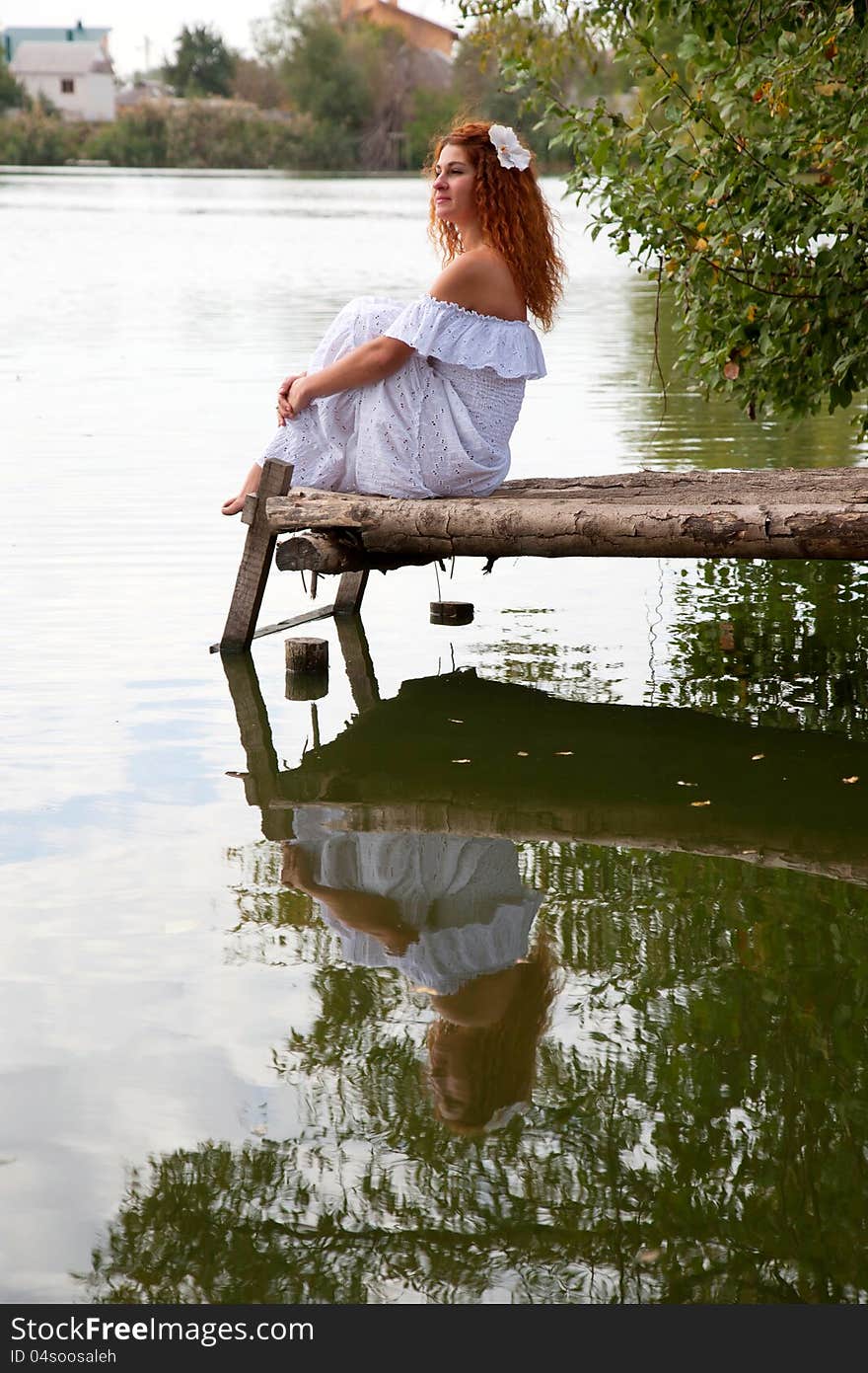 Beautiful young red-haired woman in white dress with flower in her hair or bride or undine sitting on a wooden bridge on the lake. Beautiful young red-haired woman in white dress with flower in her hair or bride or undine sitting on a wooden bridge on the lake