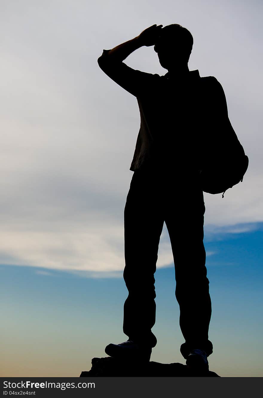 Man standing on top of a rock silhouetted against an evening sky as he stares into the distance with his hand raised to his forehead