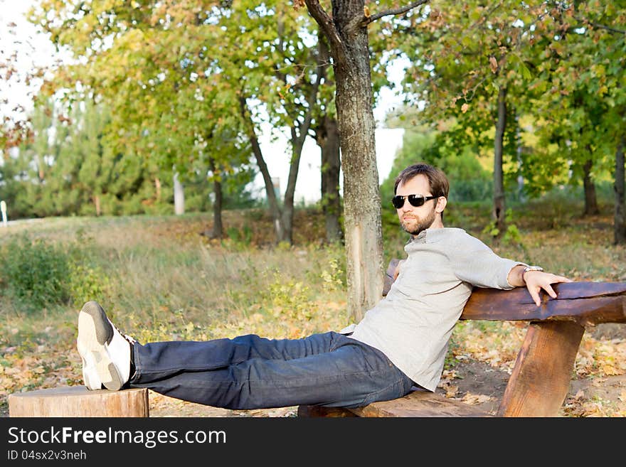 Young man relaxing in the sun sitting on a rustic wooden bench with his feet up on a tree stump with copyspace