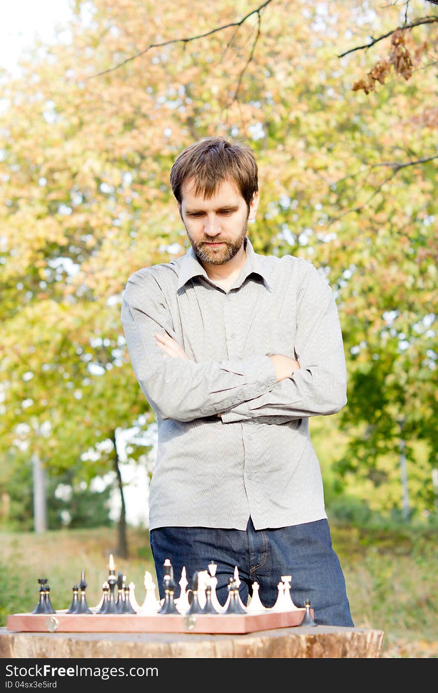 Young bearded man standing with his arms folded above an outdoor chessboard contemplating his next chess move. Young bearded man standing with his arms folded above an outdoor chessboard contemplating his next chess move