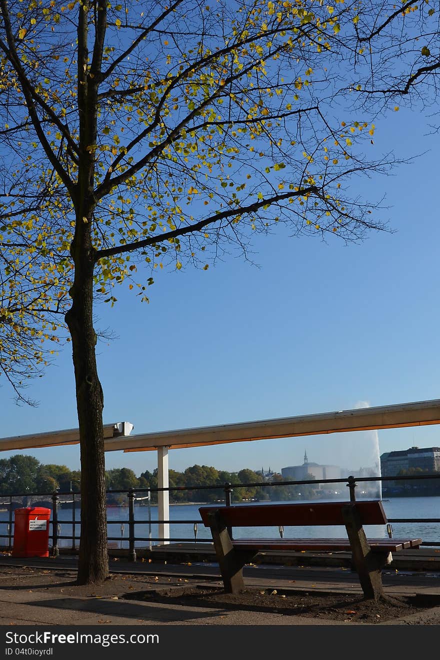 A sunny autumn day in October at the lakefront of the Binnenalster in Hamburg: Leaves are falling over the empty bench and shadows become longer. A sunny autumn day in October at the lakefront of the Binnenalster in Hamburg: Leaves are falling over the empty bench and shadows become longer.