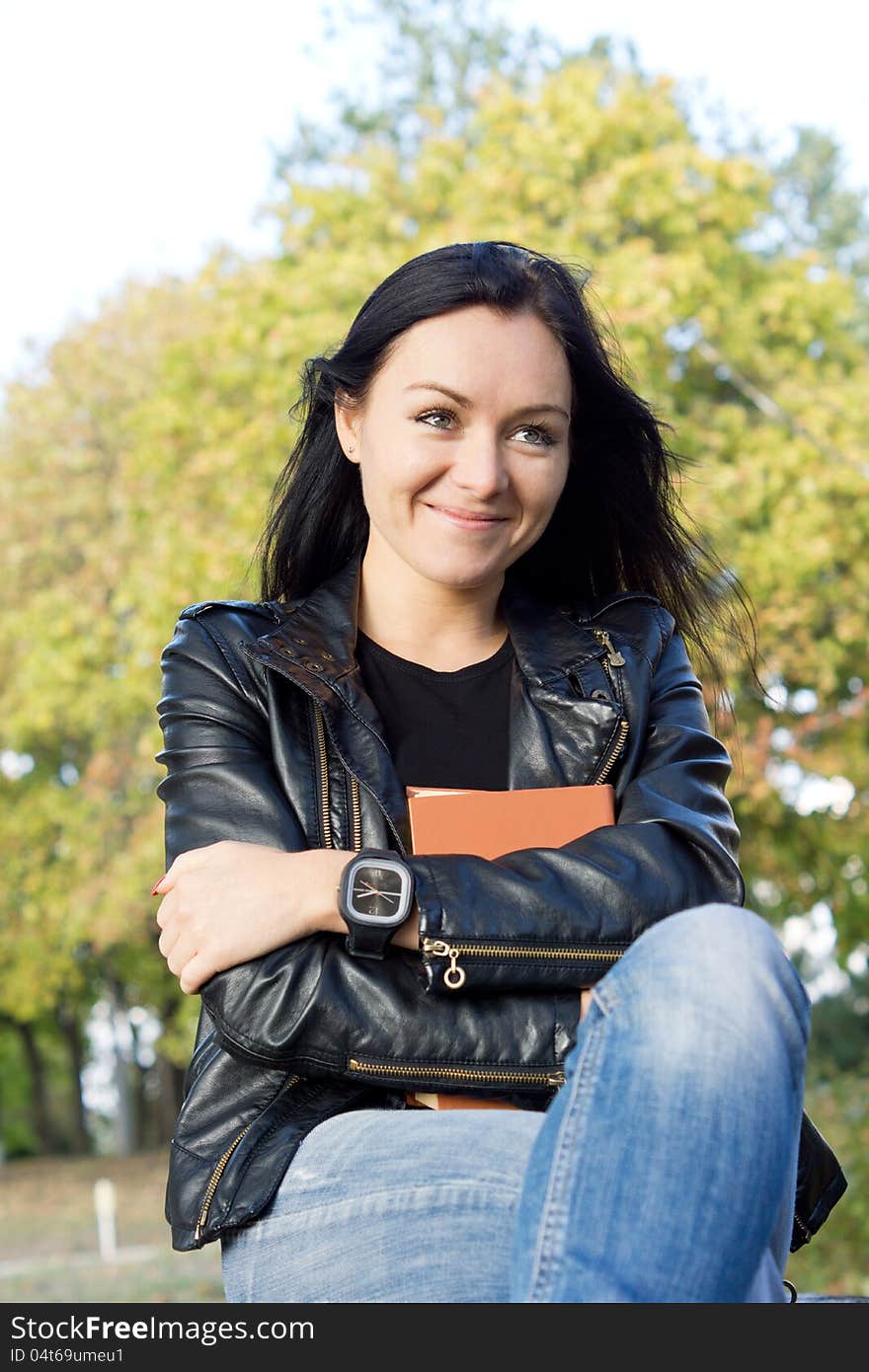 Smiling young casual womanin jeans and a leather jacket holding a book to her chest while sitting outdoors. Smiling young casual womanin jeans and a leather jacket holding a book to her chest while sitting outdoors