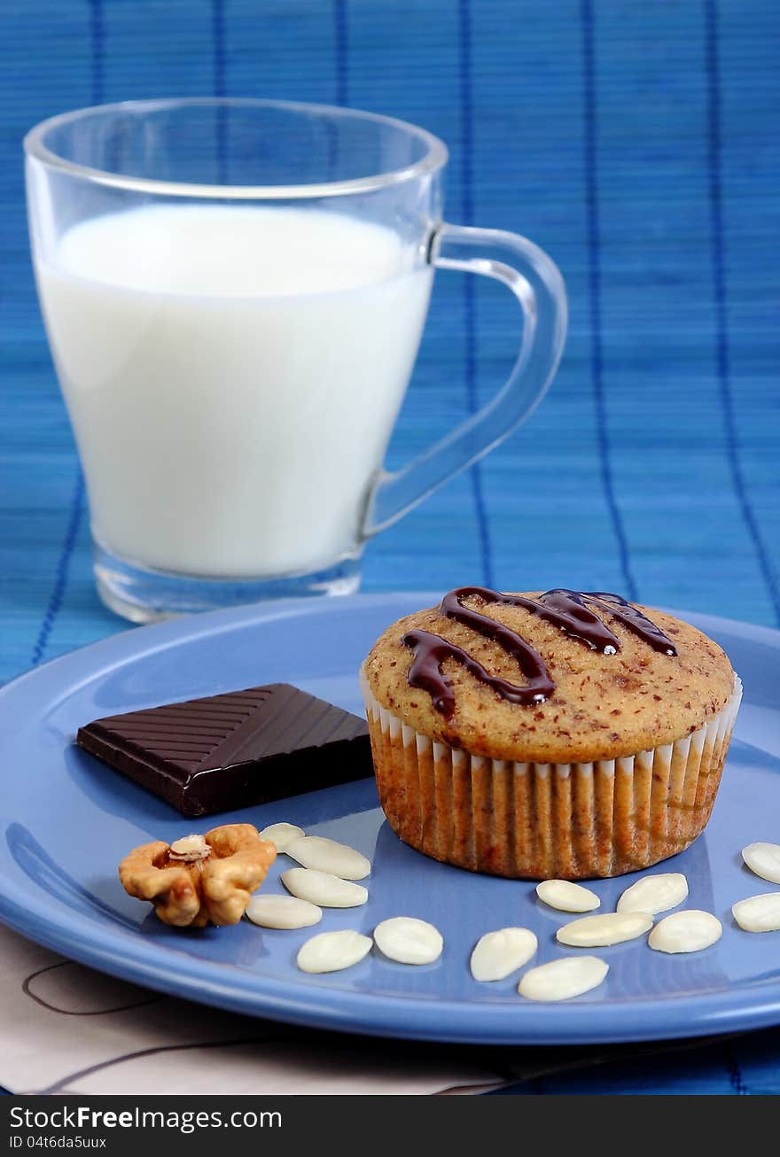 Closeup image of a muffin cake on a blue plate with almond chips, walnut, a piece if dark chocolate and a glass of milk. Closeup image of a muffin cake on a blue plate with almond chips, walnut, a piece if dark chocolate and a glass of milk