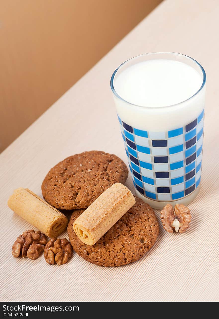 Oatmeal cookies, waffle puffs and walnuts near glass of milk decorated with blue geometric ornament on a beige wooden table in the morning. Oatmeal cookies, waffle puffs and walnuts near glass of milk decorated with blue geometric ornament on a beige wooden table in the morning