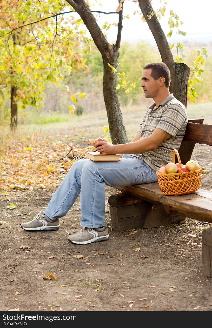 Attractive middle-aged man sitting on a rustic wooden bench outdoors eating apples with a basket full of fresh fruit at his side. Attractive middle-aged man sitting on a rustic wooden bench outdoors eating apples with a basket full of fresh fruit at his side