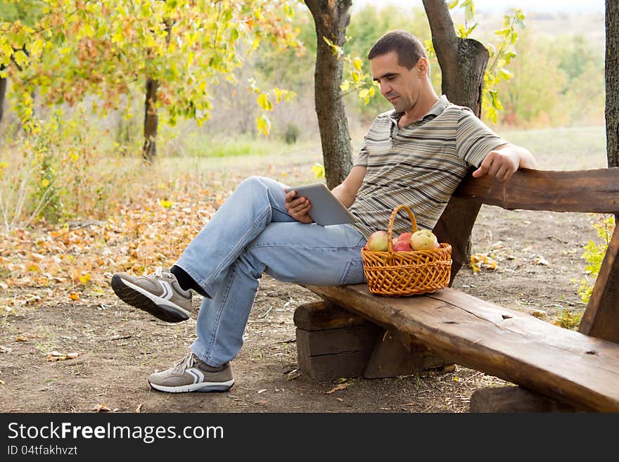 Casual middle-aged man on a rustic bench in woodland reading a book with a basket of fresh apples alongside him. Casual middle-aged man on a rustic bench in woodland reading a book with a basket of fresh apples alongside him