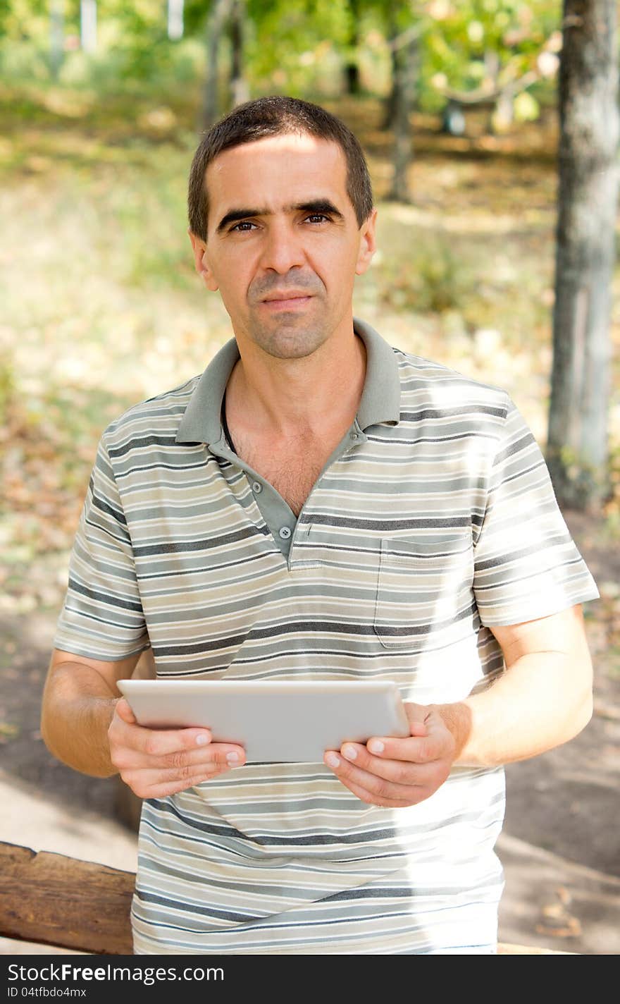 Attractive middle-aged man holding a tablet in his hands while looking at the camera, casual clothing in the garden