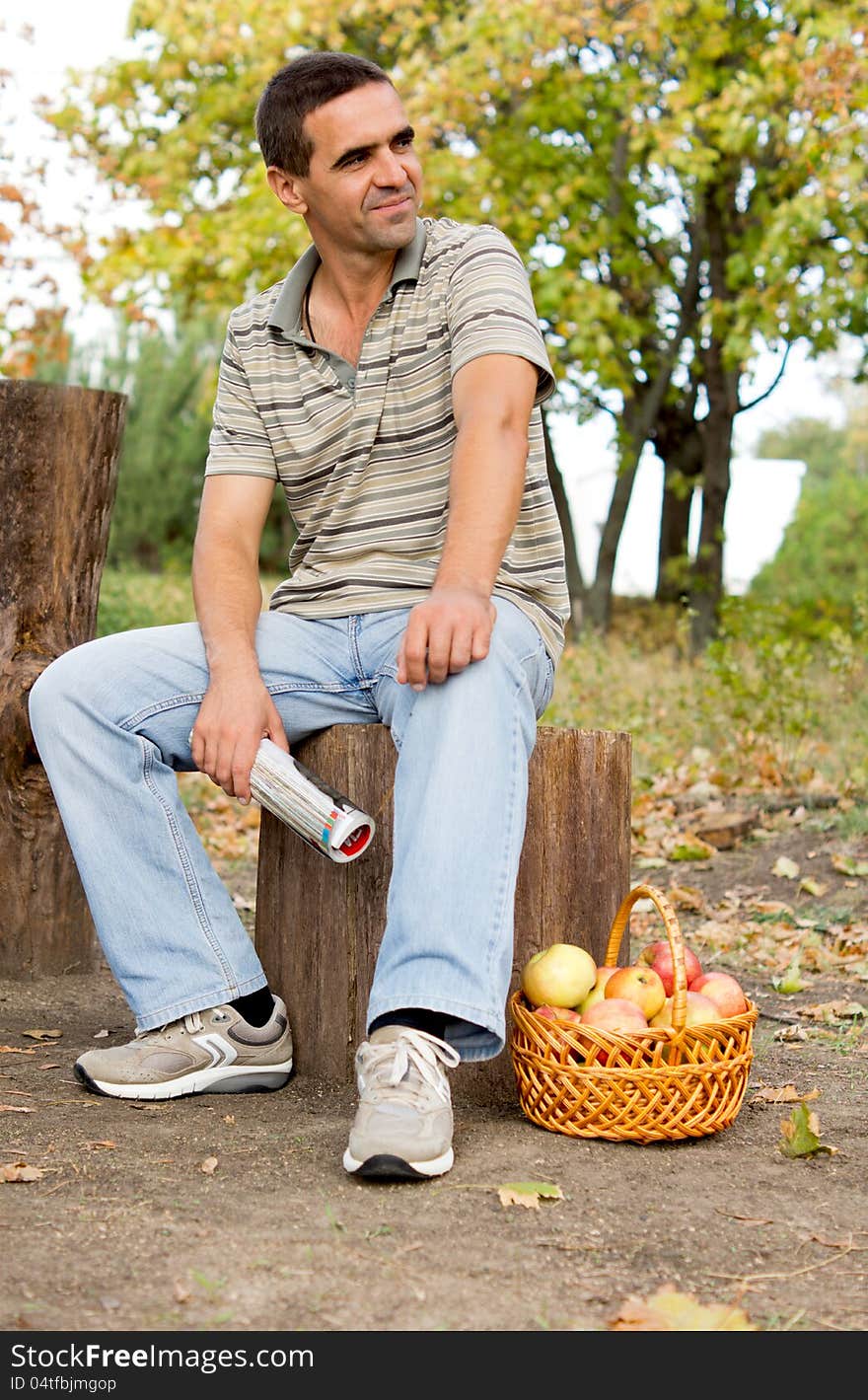 Man seated on a tree trunk in the countryside waitng patiently and looking expectantly off frame with a basket of fresh apples at his feet