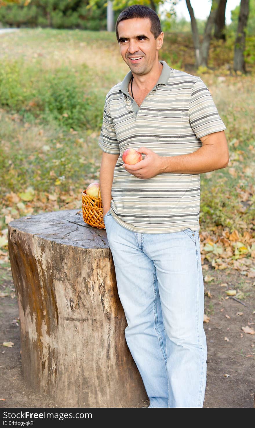 Smiling man standing alongside a sawn off tree trunk in the countryside with a fresh juicy apple in his hand. Smiling man standing alongside a sawn off tree trunk in the countryside with a fresh juicy apple in his hand