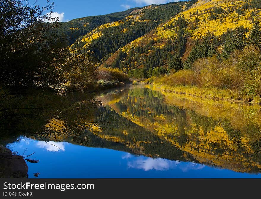 Calm mountain stream surrounded by colorful bushes and reflecting brilliant gold, yellow, and orange fall mountainside of pines, bushes, aspens and some brilliant blue sky above. Calm mountain stream surrounded by colorful bushes and reflecting brilliant gold, yellow, and orange fall mountainside of pines, bushes, aspens and some brilliant blue sky above.