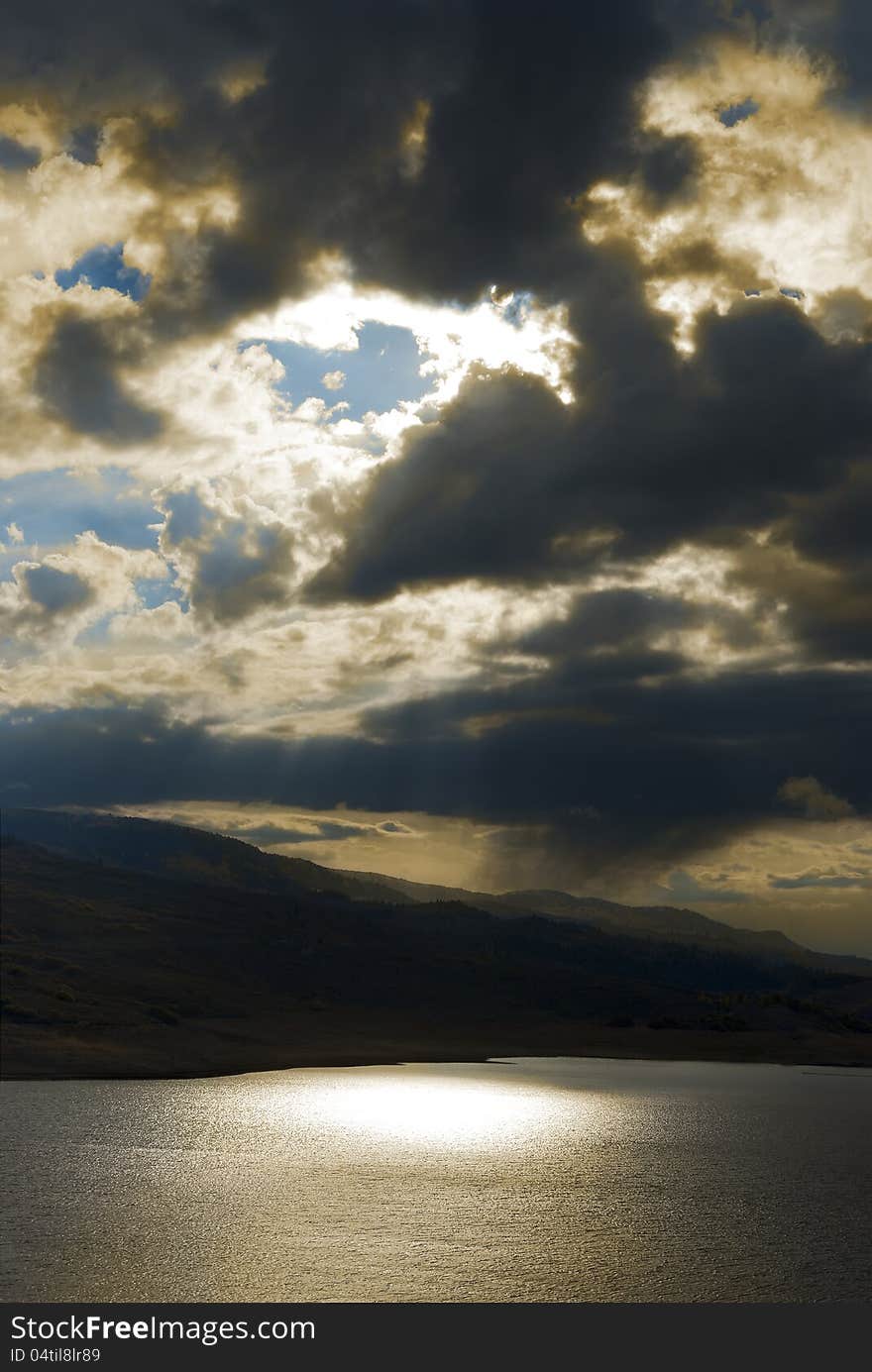 Dramatic stormy sky with sun and blue sky breaking through and light filtering through to hills and a lake glistening below. Dramatic stormy sky with sun and blue sky breaking through and light filtering through to hills and a lake glistening below.