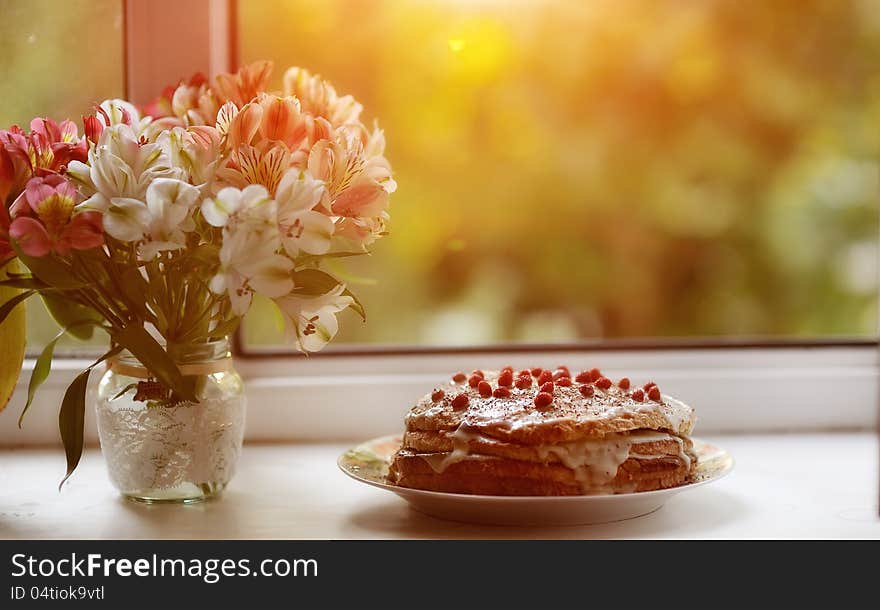 Against the background of the window is a cake with berries and a bouquet of flowers near. Against the background of the window is a cake with berries and a bouquet of flowers near