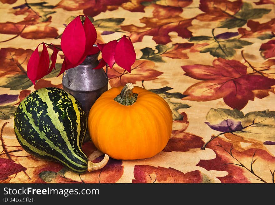 Miniature antique milk can with red leaves from a burning bush, a striped gourd, and a tiny pumpkin assembled as a centerpiece on an autumn tablecloth. Miniature antique milk can with red leaves from a burning bush, a striped gourd, and a tiny pumpkin assembled as a centerpiece on an autumn tablecloth