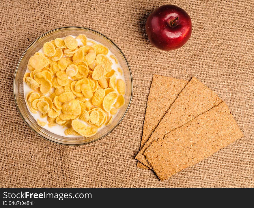 Bowl of cornflakes with milk, crispbreads and apple on sacking background