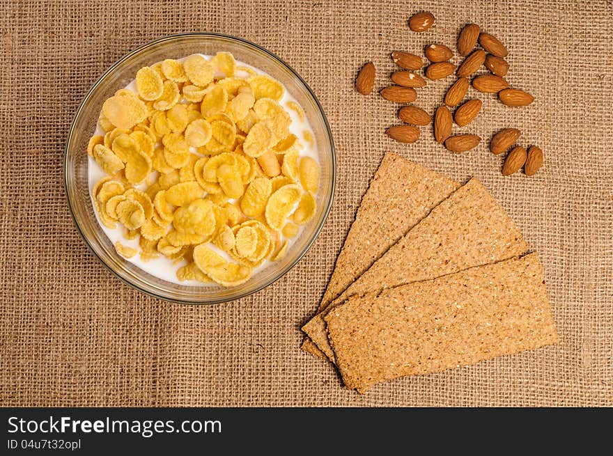 Bowl of cornflakes with milk, crispbreads and almonds on sacking background