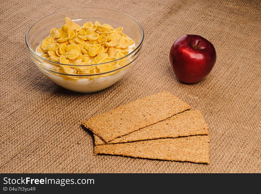 Bowl of cornflakes with milk, crispbreads and apple on sacking background