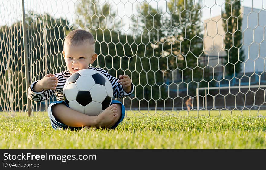 Little boy sitting cross legged on the ground in the goalposts with a soccer ball balanced on his lap. Little boy sitting cross legged on the ground in the goalposts with a soccer ball balanced on his lap