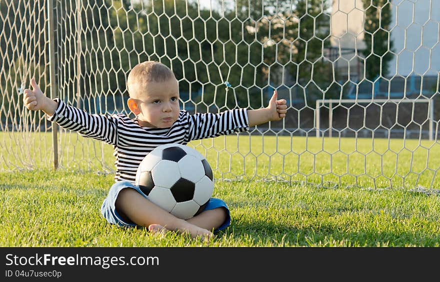 Little sportsman sitting cross lgged on green grass in the goalpost with a soccer ball on his lap giving a thumbs up. Little sportsman sitting cross lgged on green grass in the goalpost with a soccer ball on his lap giving a thumbs up