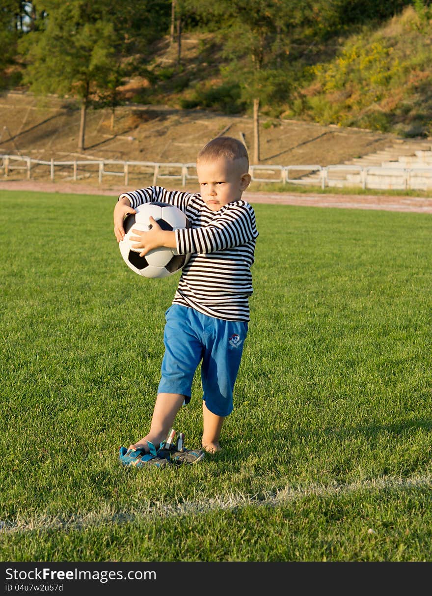 Little boy holding a soccer ball on a grassy sportsfield looking into evening sunlight with a frown on his face as he waits for someone to play with him.