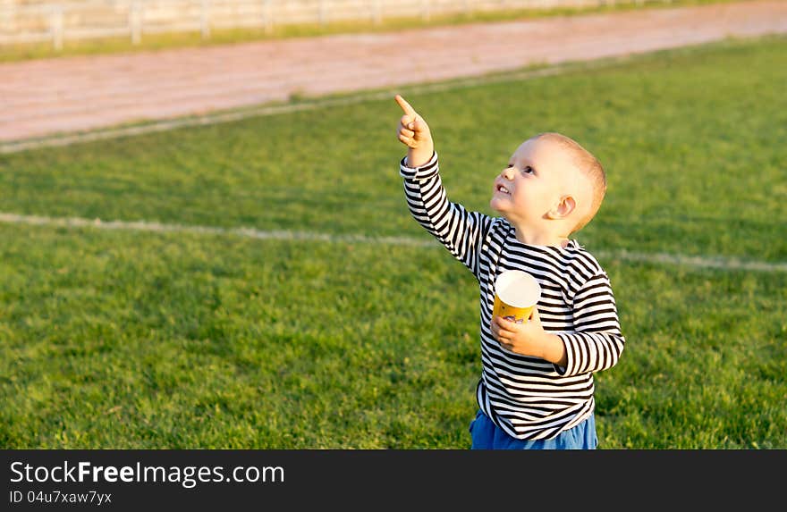 Little boy standing on a green playing field with a mug in his hand pointing at the sky. Little boy standing on a green playing field with a mug in his hand pointing at the sky